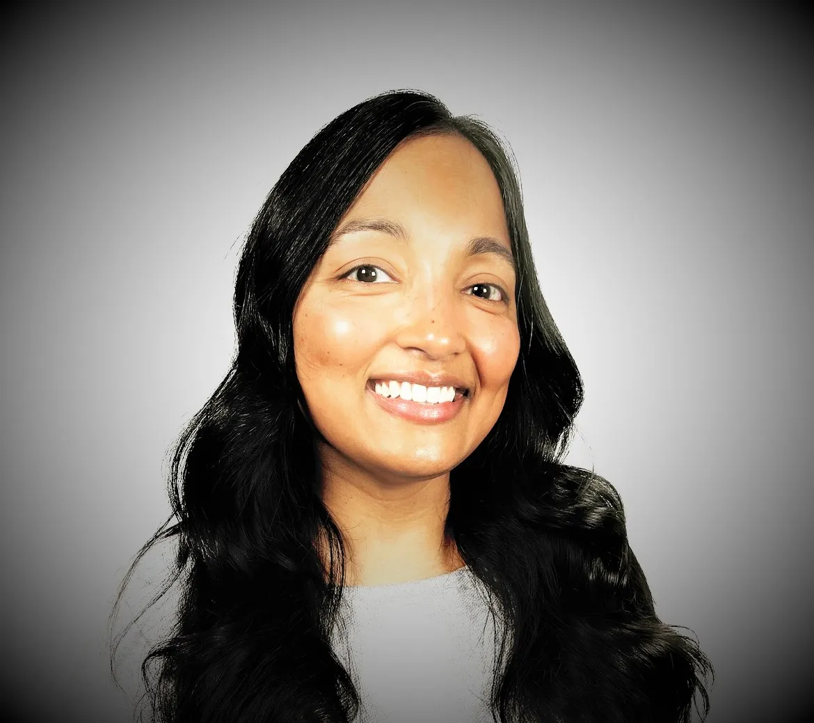 A woman with long, dark hair and a light complexion smiles at the camera against a plain background. She is wearing a light-colored top.