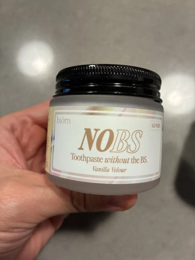 A hand holds a small jar labeled "NO BS Toothpaste without the BS. Vanilla Velour" by the brand biom, against a gray background.