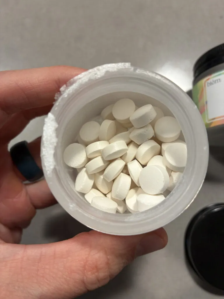 A hand holding an open bottle containing several round, white tablets on a gray surface.