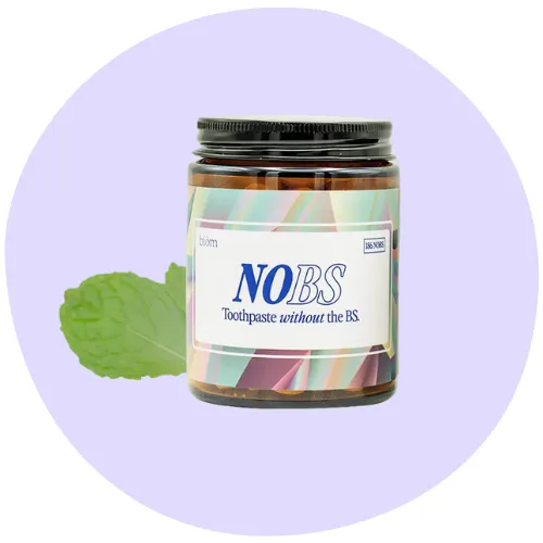 A jar labeled "NOBS Toothpaste without the BS" sits in front of a mint leaf, against a light purple circular background.