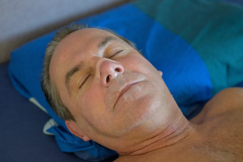 Sleeping middle aged man with a nose tape and a mouth tape lying in bed in close up