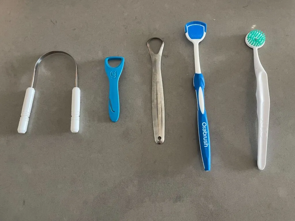 Tongues scrapers ordered from left to right (Mastermedi, Grin Tongue Cleaner, Basic Concepts, Orabrush, Tung Tongue Brush)