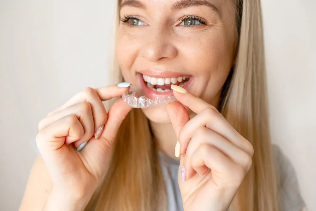 Young adult woman pictured holding Invisalign as she smiles while looking at a distance