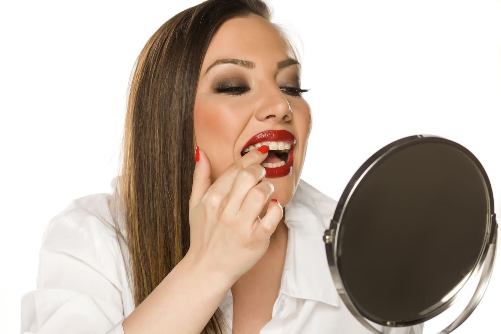 A woman in a white shirt applies red lipstick while looking into a handheld mirror against a white background.