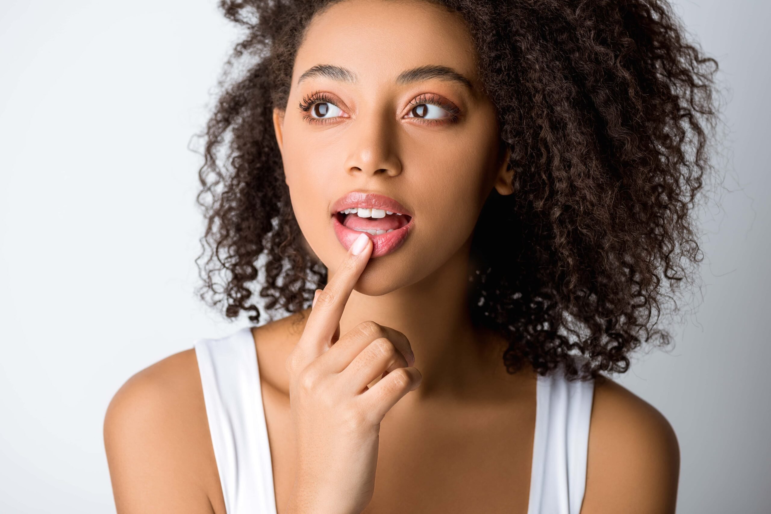 A woman with curly hair touches her lip with her finger, looking thoughtful, wearing a white top, and posing against a plain light background.