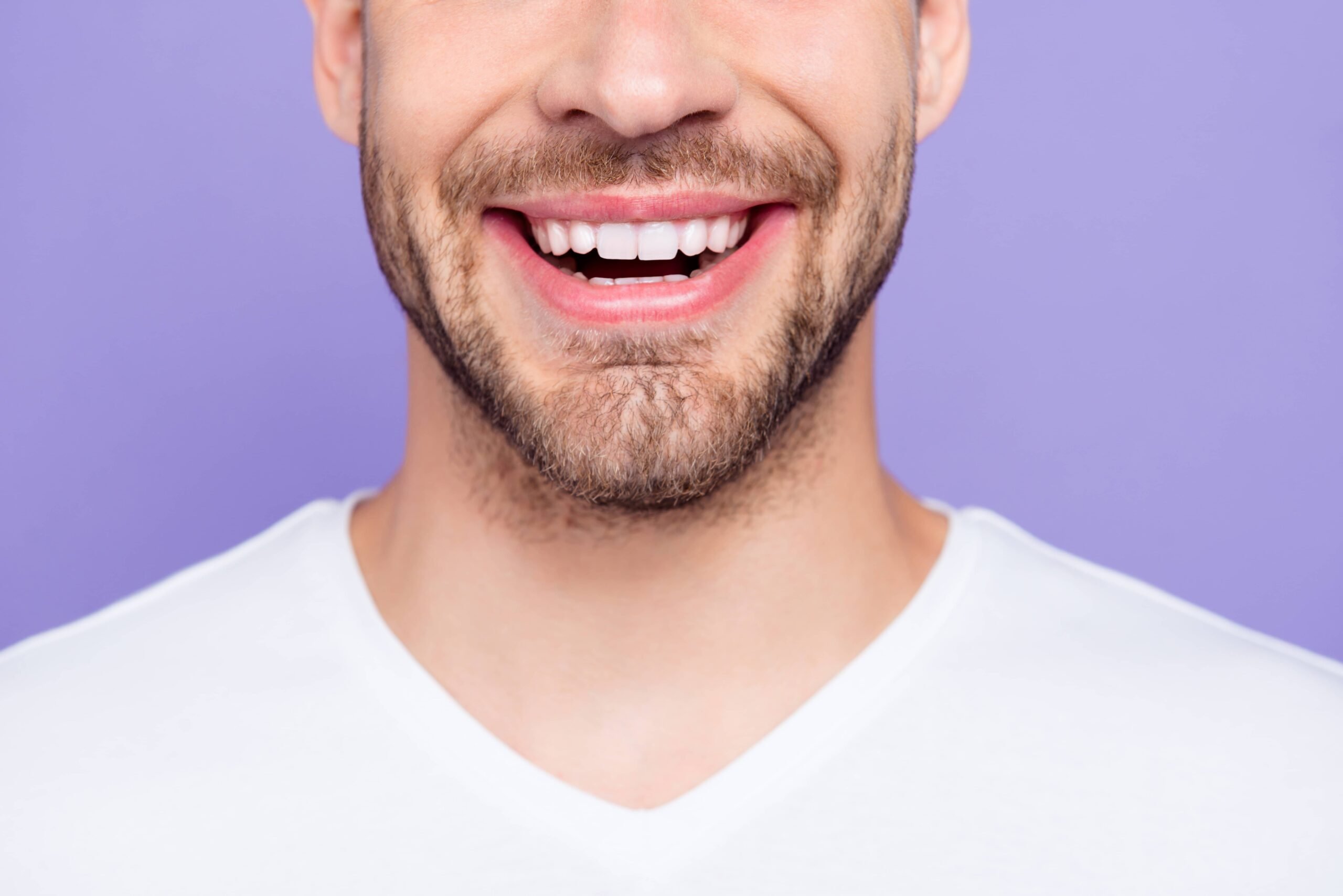 Close-up of a smiling man with a beard wearing a white shirt against a purple background. Only the lower half of his face is visible.