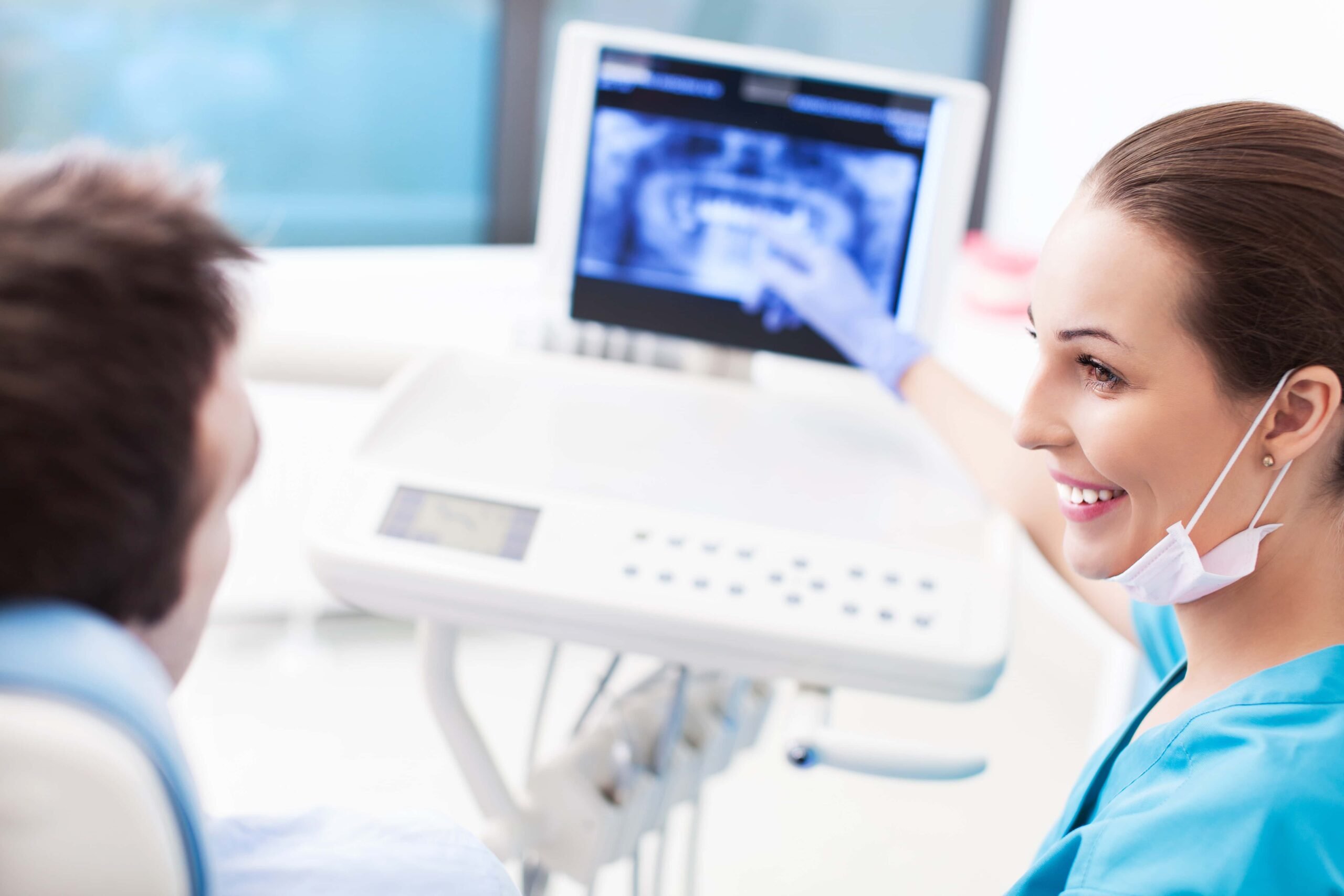A dental professional in scrubs shows a dental X-ray to a seated patient, explaining the image.