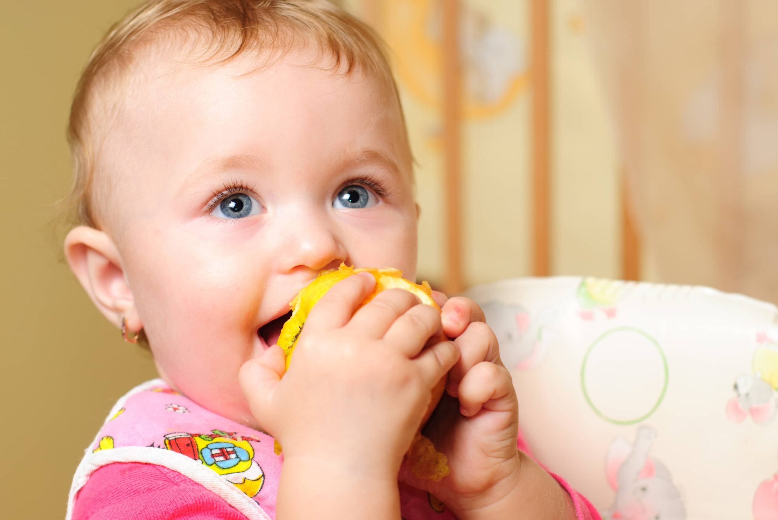 A baby with blue eyes and light hair wearing a pink bib eats a peeled orange slice, holding it with both hands. The background is softly blurred with warm tones and light patterns.