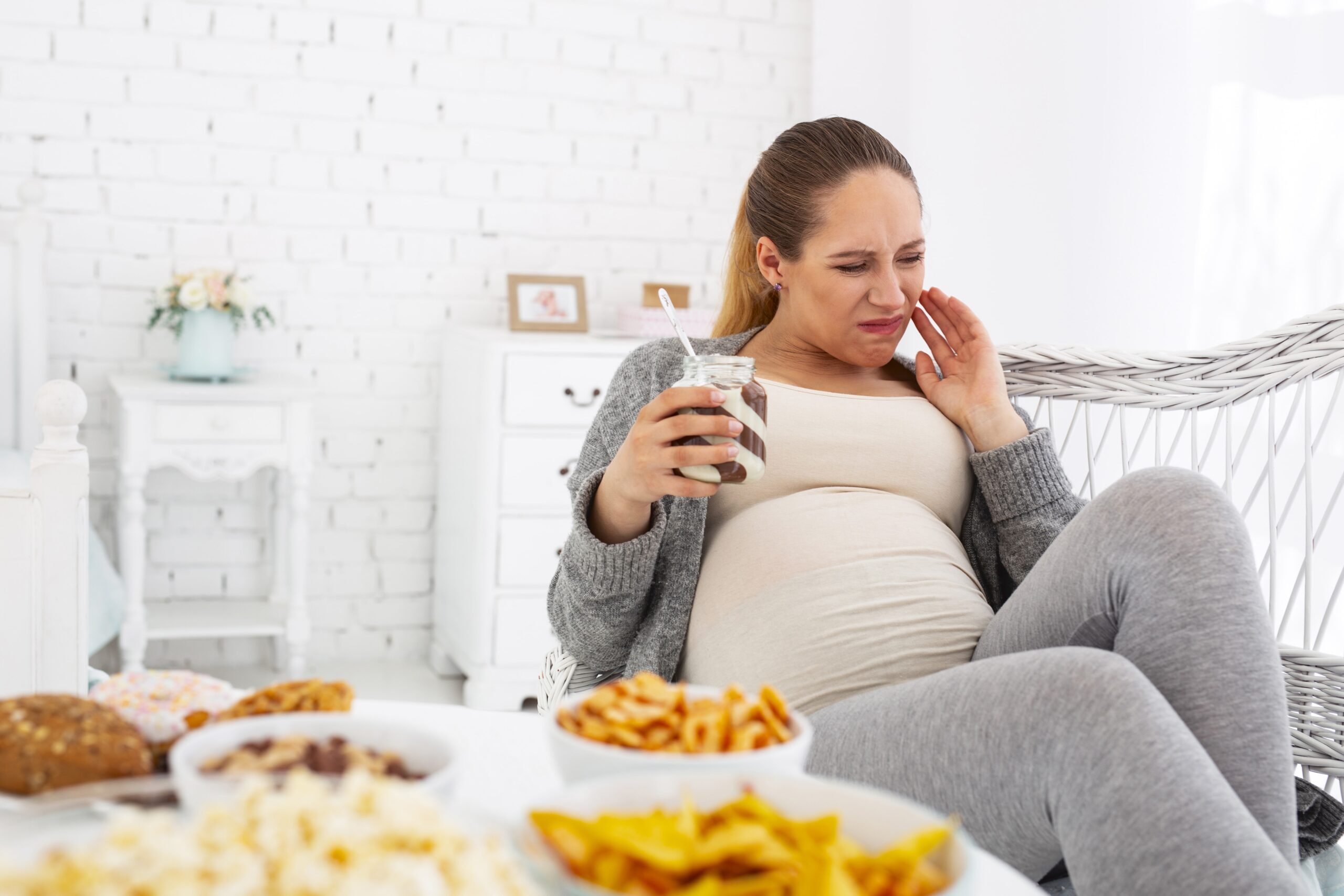 A pregnant woman in a grey sweater sits in a chair, grimacing while holding a jar of food. Various snacks are placed on the table in front of her.