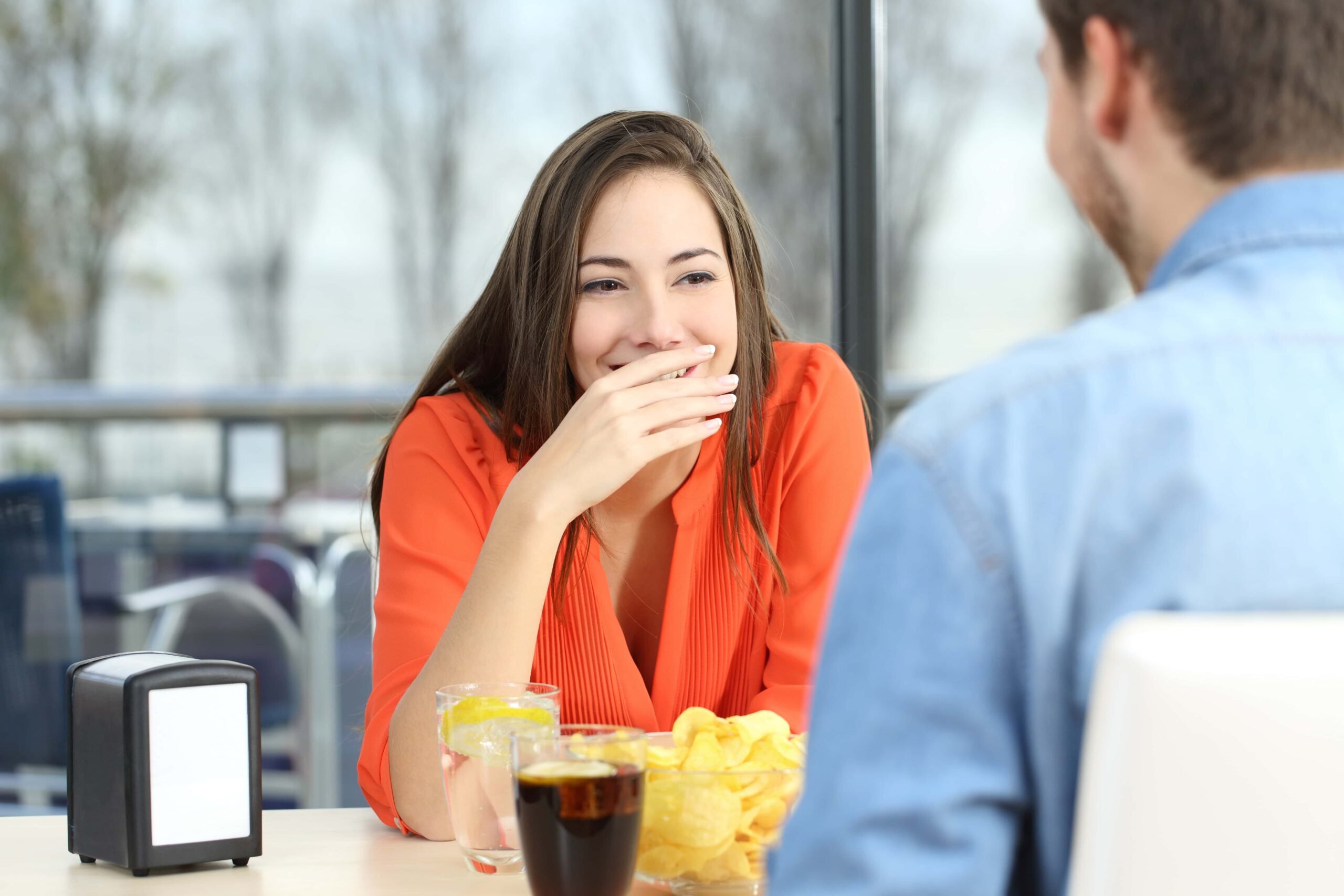 A woman wearing an orange shirt covers her mouth with her hand while smiling and sitting across from a man. They are at a table with drinks and snacks.