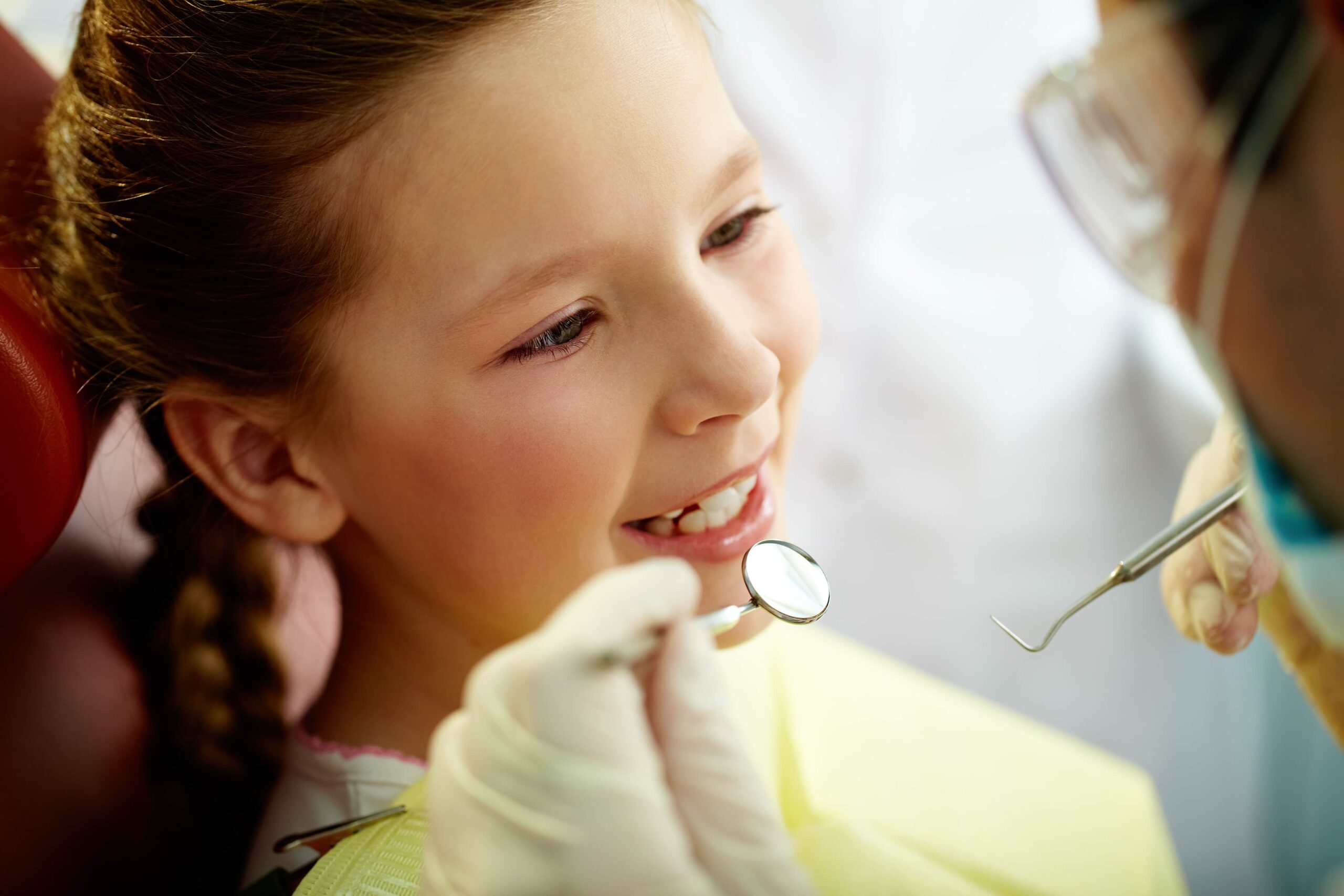 A young girl sits in a dental chair as a dentist uses a mirror and an instrument to examine her teeth.
