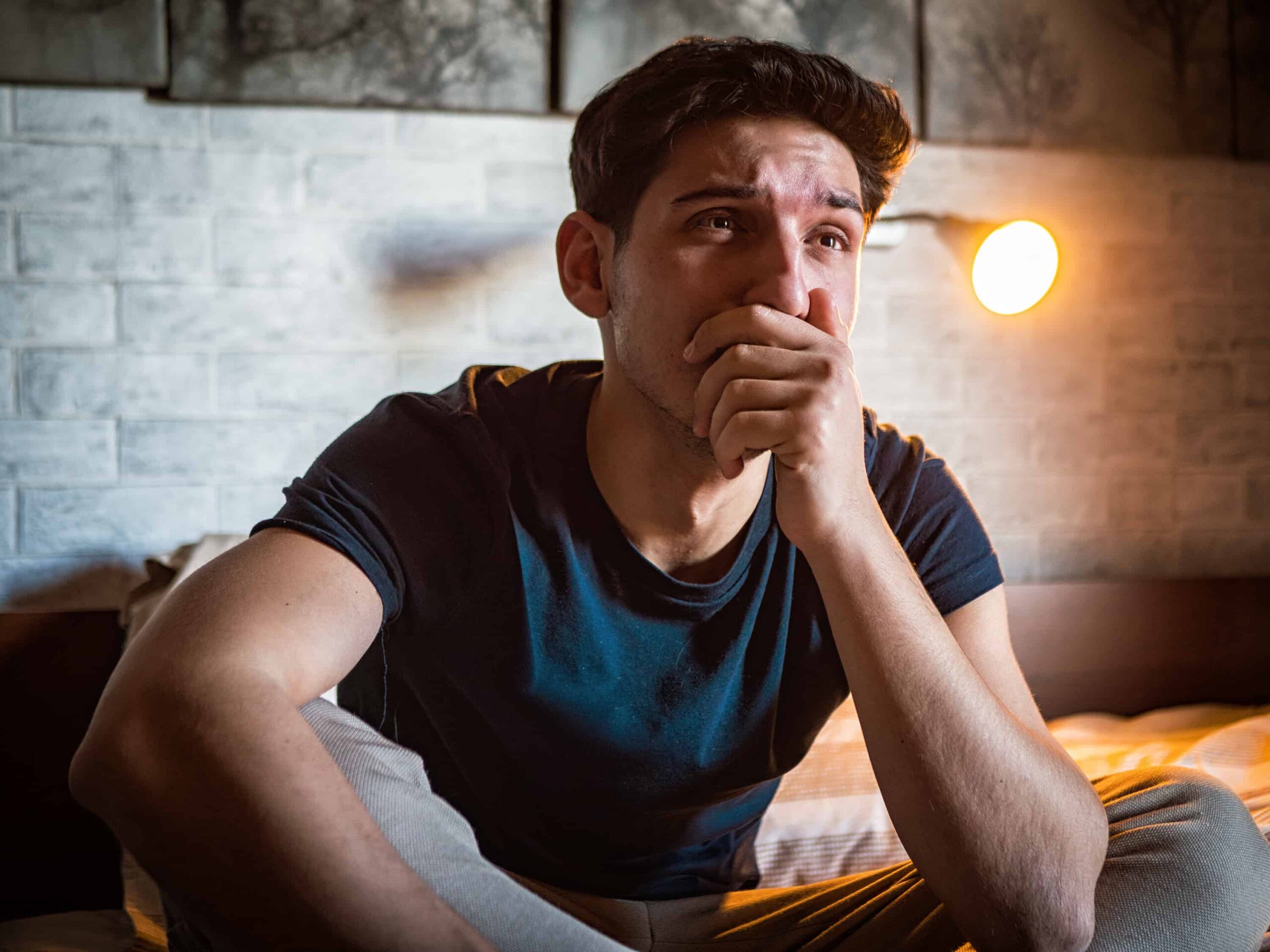 A man sits on a bed in a dimly lit room, with a hand on his face, appearing contemplative or upset.