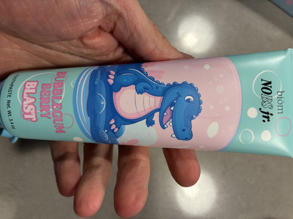 A hand holds a tube of biom NOBS jr. toothpaste, Bubblegum Berry Blast flavor, featuring a cartoon dinosaur on the packaging.