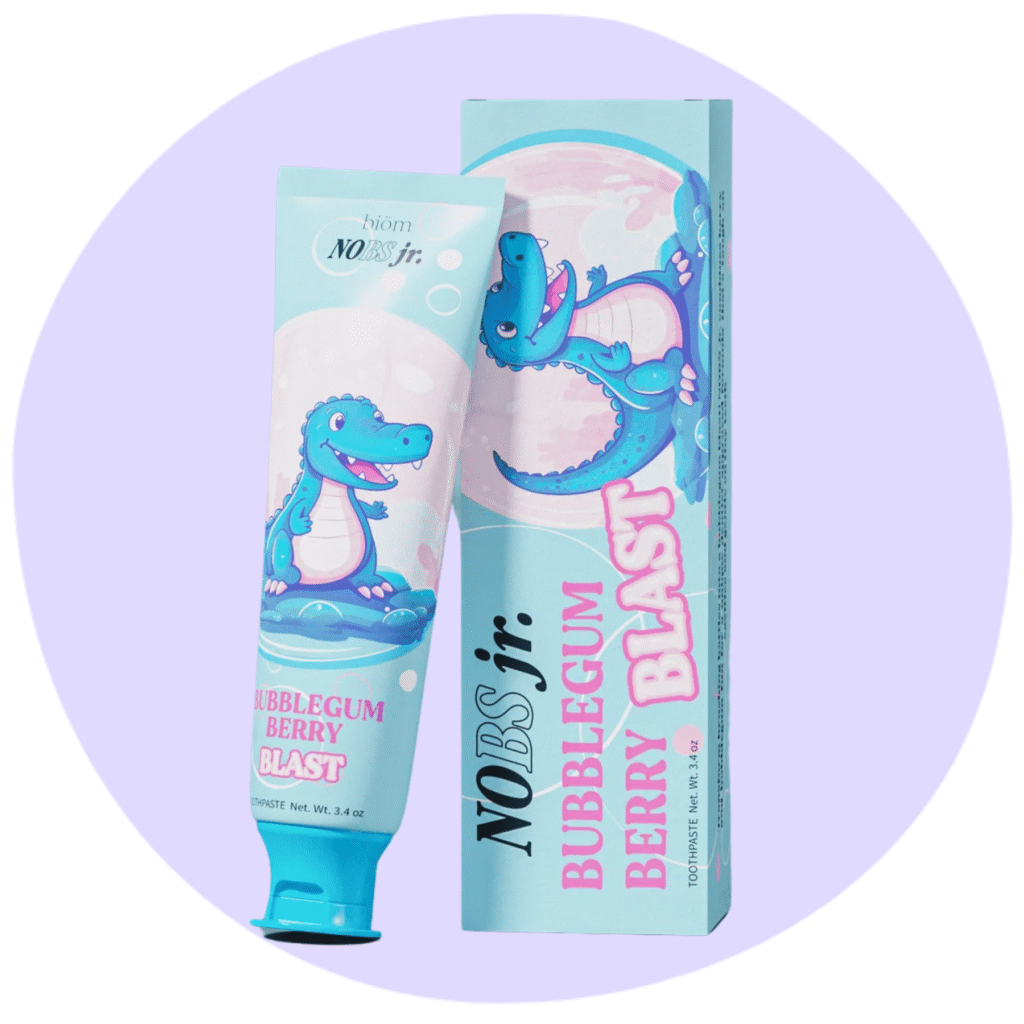 A tube and box of NOBS Jr. toothpaste in Bubblegum Berry Blast flavor, featuring a cartoon dinosaur on both the tube and packaging, set against a light purple circular background.